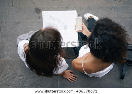 Two females are exploring a paper map and searching the information by using travel app on a mobile phone. Friends are surfing the web by using a smart-phone while traveling. View from above.