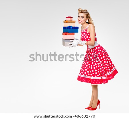 Full body of woman in pin-up style red dress in polka dot and white gloves, holding gift boxes, on grey, blank copyspace area for text, slogan. Caucasian blond model in retro fashion, vintage shoot.