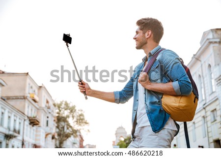 Smiling young casual man making selfie with smartphone and selfie stick at the city centre