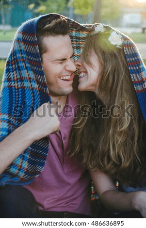 A picture of a young romantic couple covered with blanket in the park. They are laughing, kissing and man shows tongue