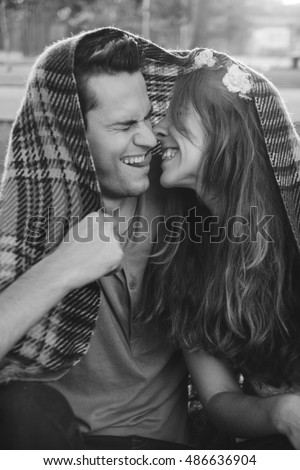 A picture of a young romantic couple covered with blanket in the park. They are laughing, kissing and man shows tongue. black and white photo