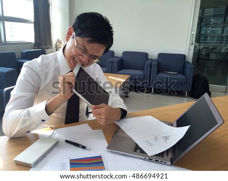 Getting serious young asian businessman trying to bite his tablet. On the table is full of data sheet, laptop and pen. The picture imply to the pressure of management in organization