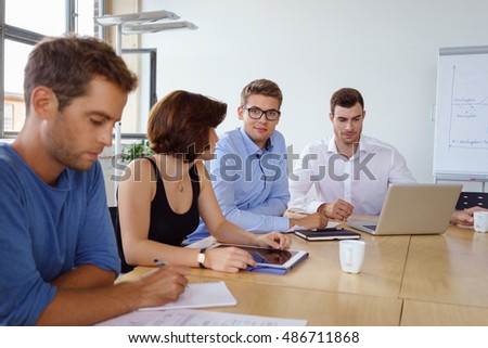 Young business team working in a boardroom seated at a conference table working on digital devices and paperwork