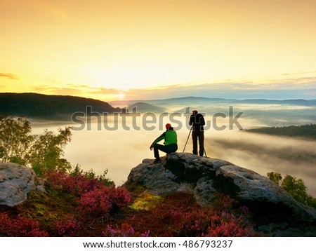 Hiker and photo enthusiast stay with tripod on cliff and takes photos. Dreamy fogy landscape, blue misty sunrise in a beautiful valley below