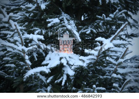 Decorative Christmas lantern with burning candle hanging on snow-covered fir-tree branch in a winter park. New year festive card, poster, postcard design. 