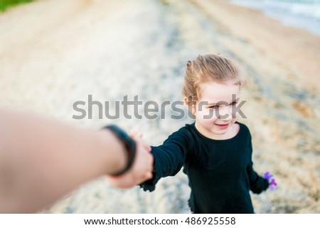Beautiful little blonde girl in a black jacket and blue jeans, walks hand in hand with parent at the sandy beach. Travel and Vacations.