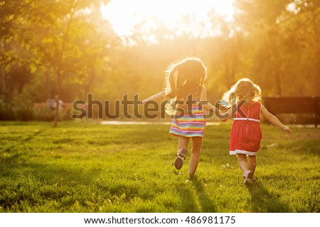 Two sisters running on the lawn in the city park outdoor. Freedom and carefree. Happy childhood. Man is unrecognizable. Royalty-Free Stock Photo #486981175