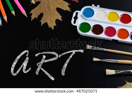 The composition of the title Art color paint, brushes, pens, pencils, paper clips, autumn leaves on a background of black school board