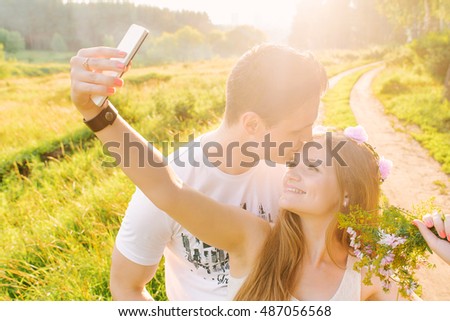 A blonde is making a selfie with her man kissing her forehead smiling happily, sunshine and green fields on the background