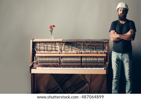 handsome bearded man with mustache and beard in building helmet or hard hat near old wooden or wood open piano with keyboard and glass vase with red rose flowers on grey background, copy space