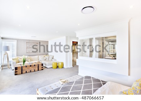 House with white ceiling and wall in the living room including a wooden box and fancy flowering plant beside the sofa and pillows, the stand lamp near to the window, there is a bed and a washroom