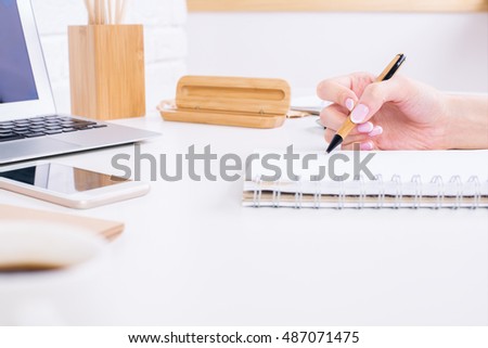 Close up and side view of woman's hand writing in spiral notepad placed on white office desktop with smartphone, laptop computer and other items