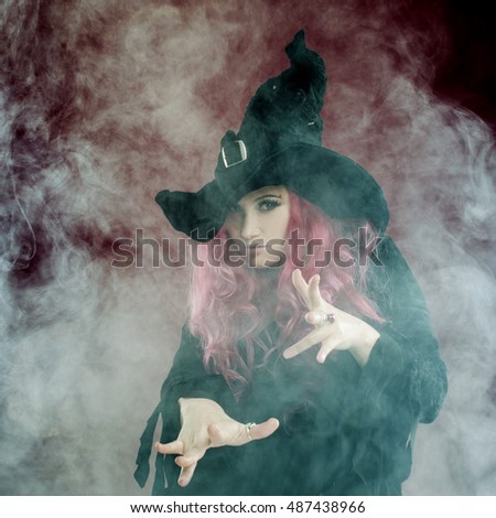 Attractive woman in witches hat and costume with red hair performs magic on a pink background. Halloween, horror theme.