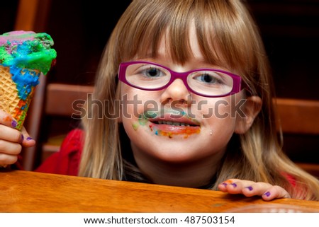 A little girl with a messy and colorful ice cream cone smiles at the camera with a messy face.  She has glasses and purple finger nails.