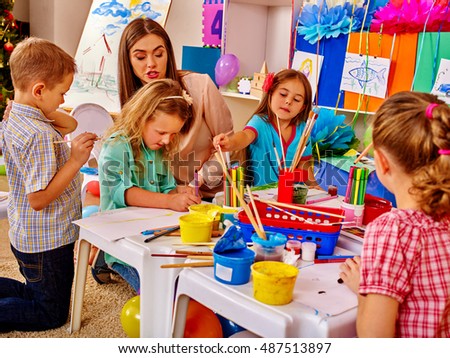 Children with teacher woman painting school on paper at table in kindergarten . Interior of kindergarten or primary school.