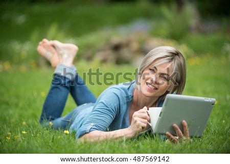 nice woman of forty years old with grey hair. she is lying in the grass , in her garden. she is looking for a journey on her tablet. she is drinking her tea. the light is on her. 