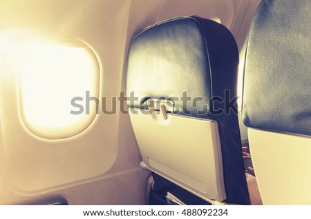 Passenger plane interior fragment. Main cabin chairs with folding tables and glowing porthole with natural sunlight lens flare. Vintage tonal correction filter, warm old style photo effect