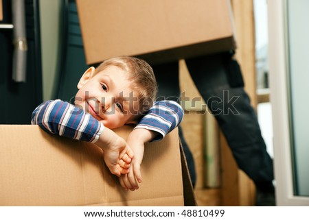 Family moving in their new home. The son is sitting inside a moving box. In the background the father - or a mover (only legs to be seen) is carrying boxes inside the building