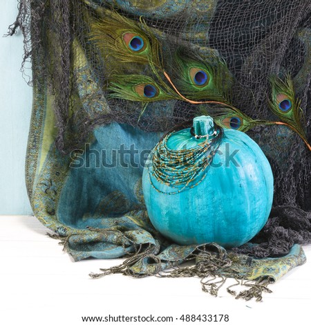 Exotic BOHO style Halloween display with a teal pumpkin, seed bead necklace, peacock feathers, netting and fringed fabric in blue and green jewel tones in square format