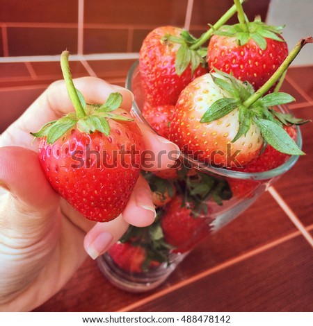 close up woman hand holding fresh strawberry with many strawberry in a glass at background