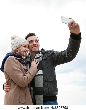 Portrait of tourist couple taking selfies together using smart phone, networking on winter beach holiday, travel lifestyle. Boyfriend and girlfriend hugging taking pictures using technology outdoors.