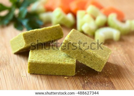 Vegetable bouillon, stock or broth cubes with parsley and fresh celery and carrot in the back, photographed with natural light (Selective Focus, Focus on the upper edge of the two cubes in the front)