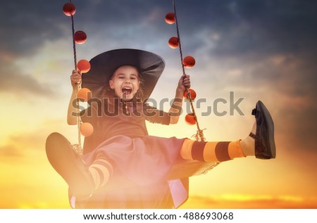 Happy Halloween! Cute little witch on swing in sunset. Beautiful young child girl in witch costume outdoors. 