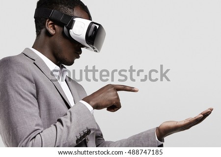 Dark-skinned man in formal wear with virtual reality headset for smart phone on his head, playing video games., holding his hands as if typing message on electronic device, looking concentrated