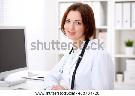 Young brunette female doctor sitting at a desk and working on the computer at the hospital office.  Health care, insurance and help concept. Physician ready to examine patient