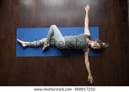 Attractive young woman working out indoors, doing yoga exercise on wooden floor, lying in Reclining Spinal Twist, Jathara Parivartanasana, resting after practice, full length, top view Royalty-Free Stock Photo #488817250