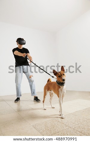 A young model in VR glasses, jeans and blank black t-shirt holding a basenji dog on a leash