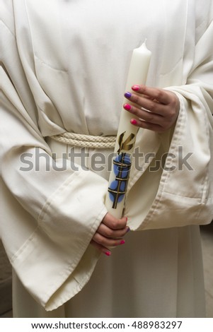 A young girl with colorfully painted finger nails wearing a white robe and carrying a big candle during a catholic celebration.