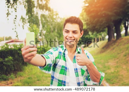 Young handsome brunet mixed race millennial man taking a selfie on smart phone, gesturing thumbs up, outdoors in autumn. Vibrant colors, natural light.