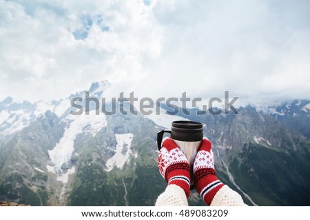 Closeup photo of thermos mug with tea in traveler's hand over out of focus mountains view with snow, tourizm in cold season