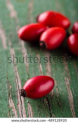 Vertical photo of several red hips with one placed aside. Group of autumn fruit is placed on wooden board with worn green color and nice texture of surface. Fruit is harvested from bush.