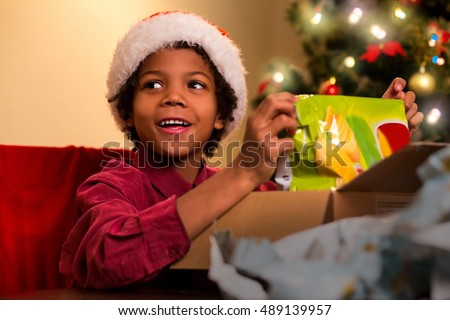 Smiling darkskinned boy unpacks present. Afro kid opens Christmas present. Happiest moment of evening. Holiday for every child.