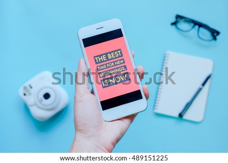 Hand holding smartphone and inspirational quote on screen with camera, notebook and eyeglasses on blue background