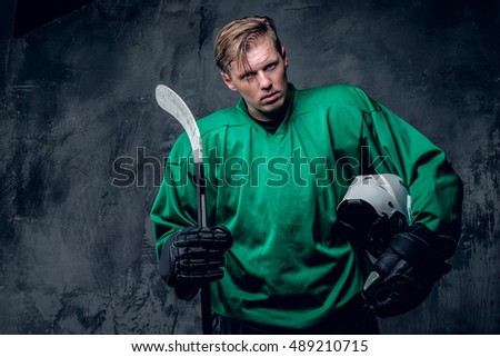 The blond Caucasian hockey player holds protective helmet and playing stick on grey background.