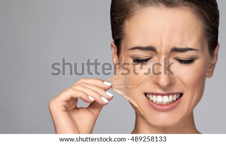 Portrait of brunette young Beautiful woman in facial peel off mask. Peeling. Beauty girl and body skin care. Isolated on grey background. Studio shot. Pull piece of leather to the side. Hand holding Royalty-Free Stock Photo #489258133