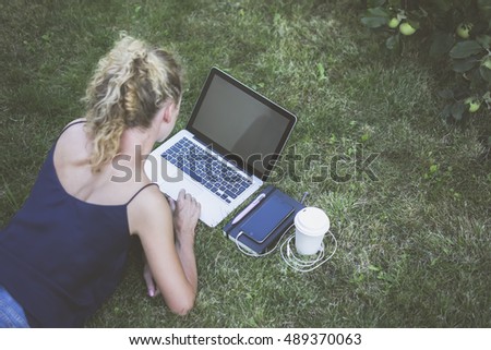 Summer,rear view and top view,girl with blond curly hair lying on grass and using laptop with blank screen.Next to computer is black notebook,pen,smartphone,wired headphones and a white coffee cup.