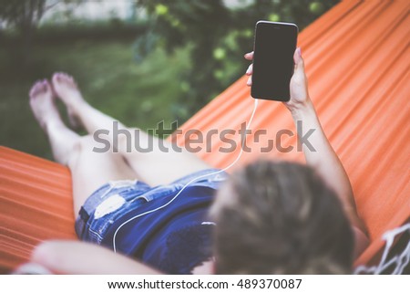 View from behind a young woman lying in a hammock in the park and using a smartphone.Close-up of a smartphone with a blank screen in the hands of a girl lying in the orange hammock.Mock up.Film effect