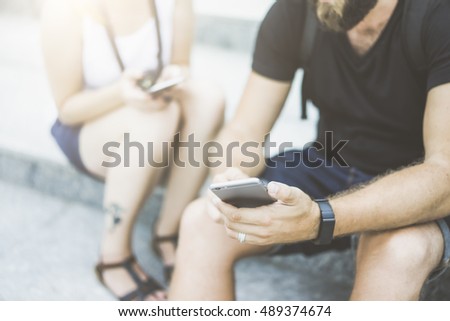 Closeup of a smartphone in male hands.Man in black T-shirt and denim shorts sitting outside on steps and using digital gadget.In the background sits blurred woman with a phone in his hand.Film effect.