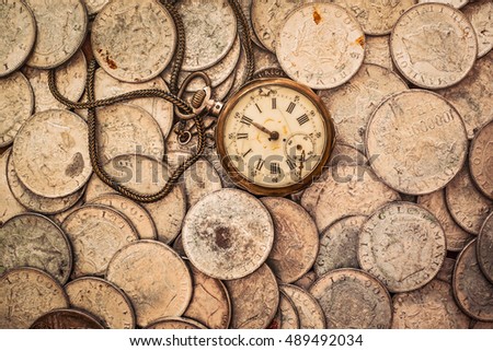A bunch of coins with a broken pocket watch on top, top view.