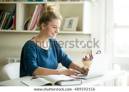 Portrait of young beautiful happy smiling casual woman working or studying using laptop computer at small home office or in the student dorm. Indoors image Royalty-Free Stock Photo #489592156