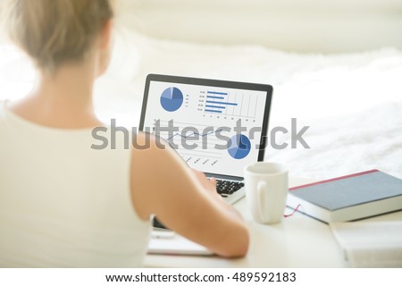 Close-up rear view of young business or student woman working at home using laptop computer, typing, looking at screen with statistic. View over the shoulder. Modern workplace, focus on monitor