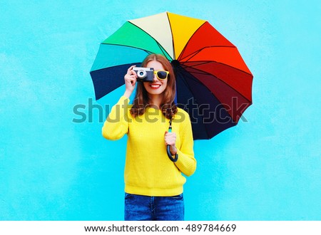 Happy smiling young woman with vintage camera holding colorful umbrella in autumn day over blue background wearing yellow knitted sweater