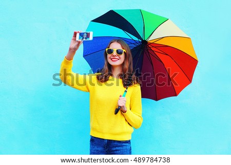 Fashion pretty smiling woman with colorful umbrella taking autumn photo makes self portrait on smartphone over blue background wearing a yellow knitted sweater