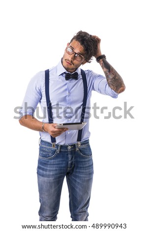 Portrait of brunette young man in light blue shirt, suspenders and jeans, using tablet PC, standing in studio shot isolated against white background.