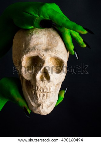 Green hand with black nails holding a human skull. On a black background.