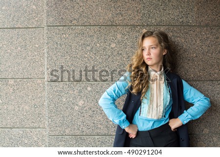 A teenager girl  (Caucasian white) (blonde) with beautiful curls in a blue blouse, a dark jacket and trousers demonstrates a business clothes style in streets of a city. 